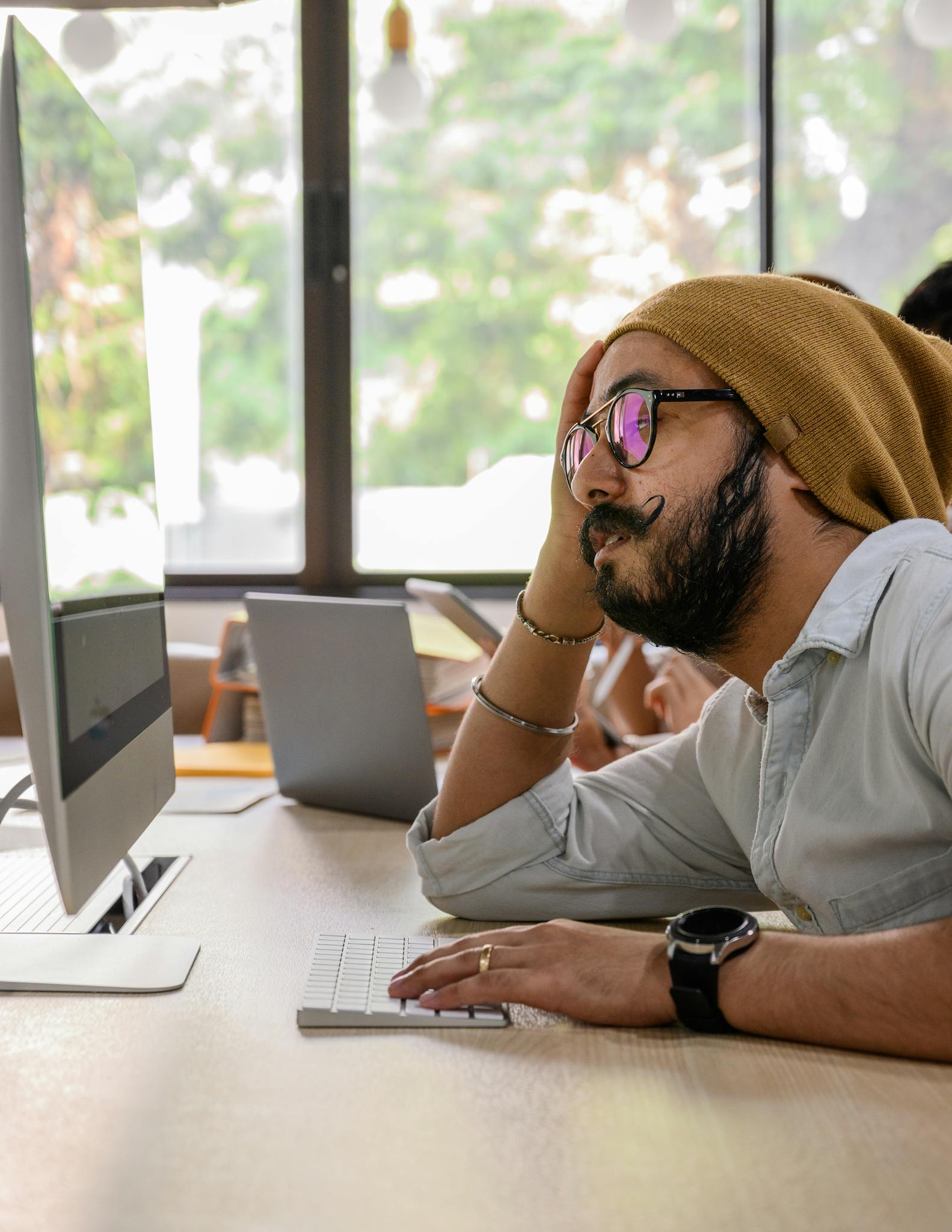 A man with glasses and hat working on a computer in a modern office with natural light.